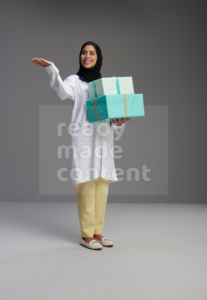Saudi woman wearing lab coat with stethoscope standing holding gift box on Gray background