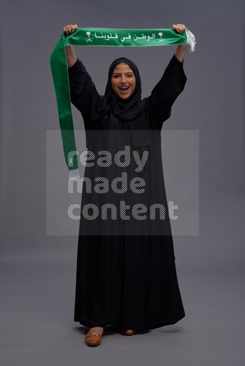 Saudi woman wearing Abaya standing holding Saudi flag on gray background
