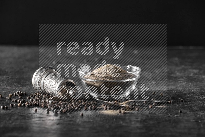 A glass bowl full of black pepper powder with black pepper beads, a turkish metal pepper grinder and a metal spoon on textured black flooring