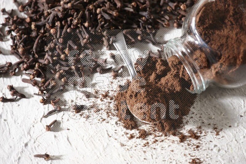 وعاء زجاجي مقلوب ممتلئ ببودرة القرنفل وتناثرت البودرة منه مع حبوب القرنفل المنتشرة على أرضية بيضاء