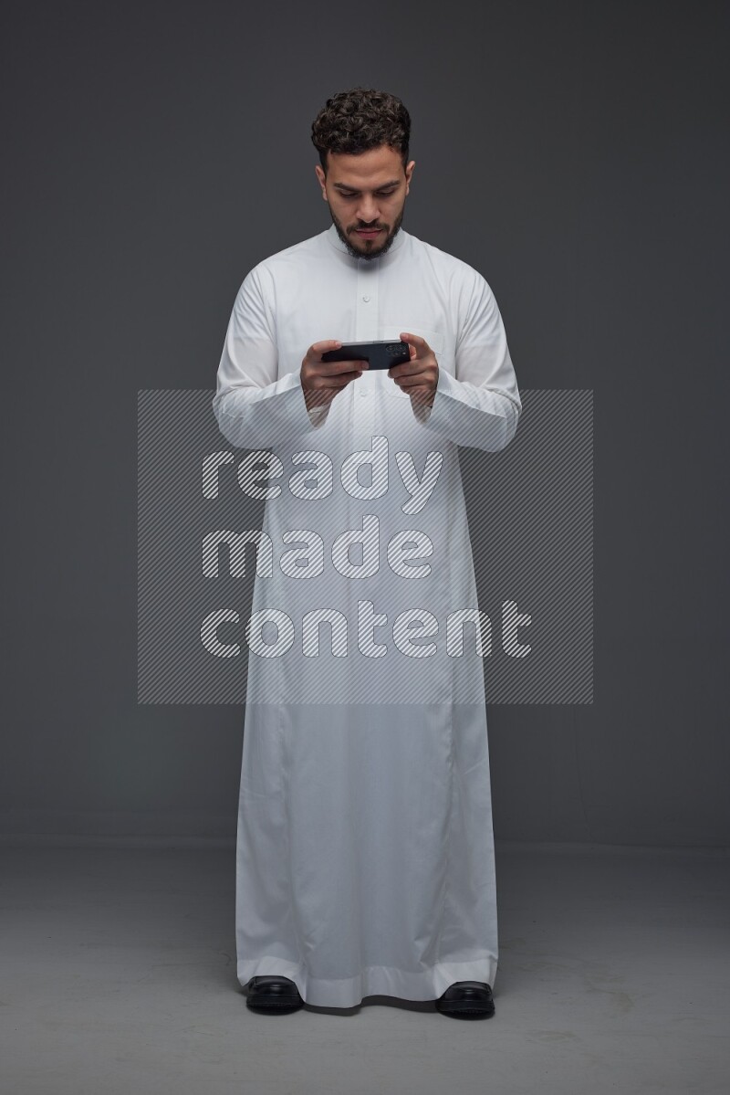A Saudi man wearing Thobe and playing using his phone horizontally playing video games while standing and making different poses eye level on a gray background