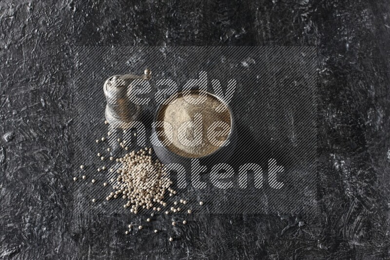 Black pottery bowl full of white pepper powder with pepper beads and metal pepper grinder on textured black flooring