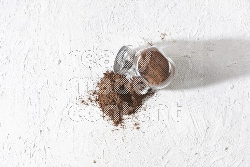A flipped glass spice jar full of cloves powder and powder came out of it on textured white flooring