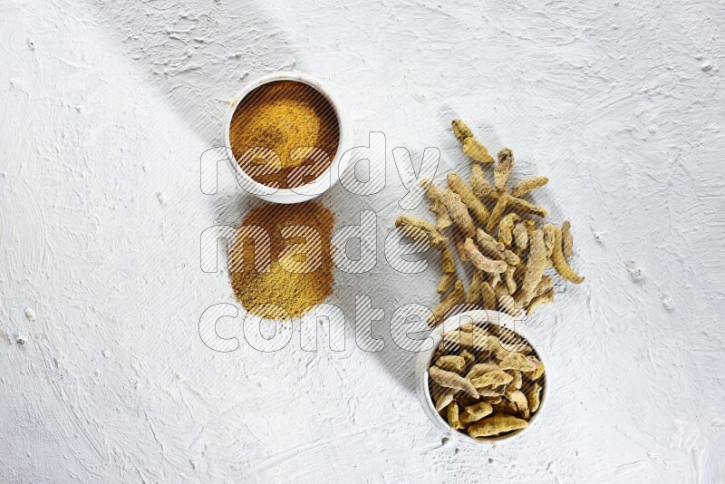 وعاءان فخاريان بيج ممتلئان ببودرة الكركم وأصابع الكركم المجففة، مع بودرة وأصابع الكركم بجانبهما على خلفية بيضاء