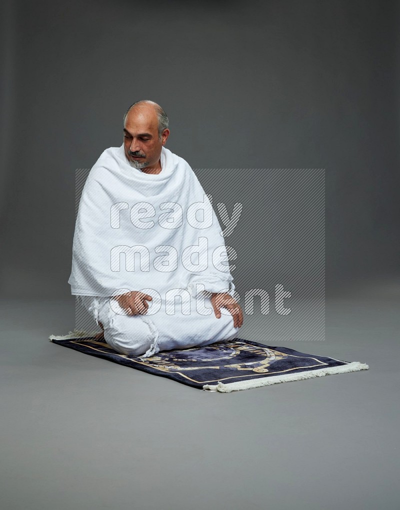 A man wearing Ehram sitting on prayer mat on gray background