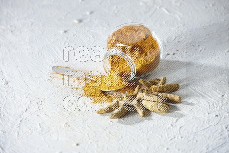 A flipped glass spice jar and metal spoon full of turmeric powder and powder spilled out of it with dried whole fingers on textured white flooring