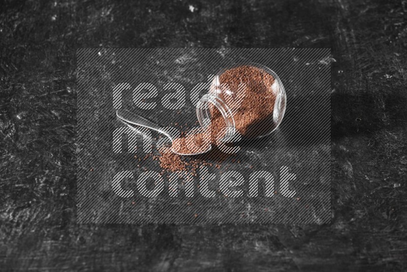 A glass spice jar full of garden cress seeds flipped and seeds spread out with a metal spoon full of the seeds on a textured black flooring