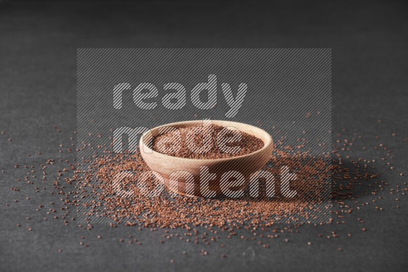 A wooden bowl full of garden cress seeds surrounded by seeds on a black flooring