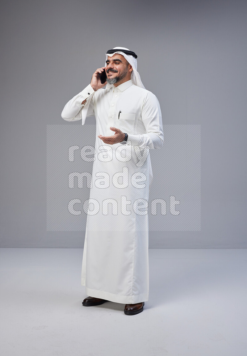 Saudi man Wearing Thob and white Shomag standing talking on phone on Gray background