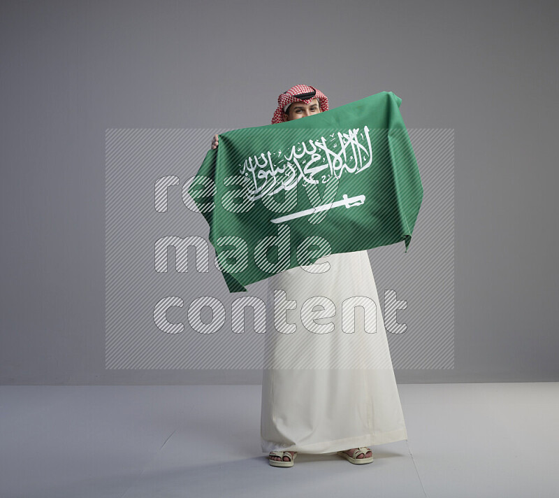 A saudi man standing wearing thob and red shomag holding big saudi flag on gray background