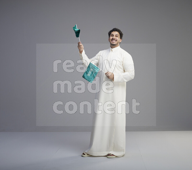 A Saudi man standing wearing thob holding small Saudi flag on gray background