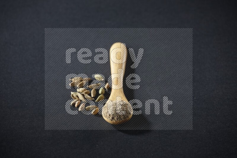 ملعقة خشبية ممتلئة ببودرة الهيل وحبوب الهيل بجانبها على خلفية سوداء