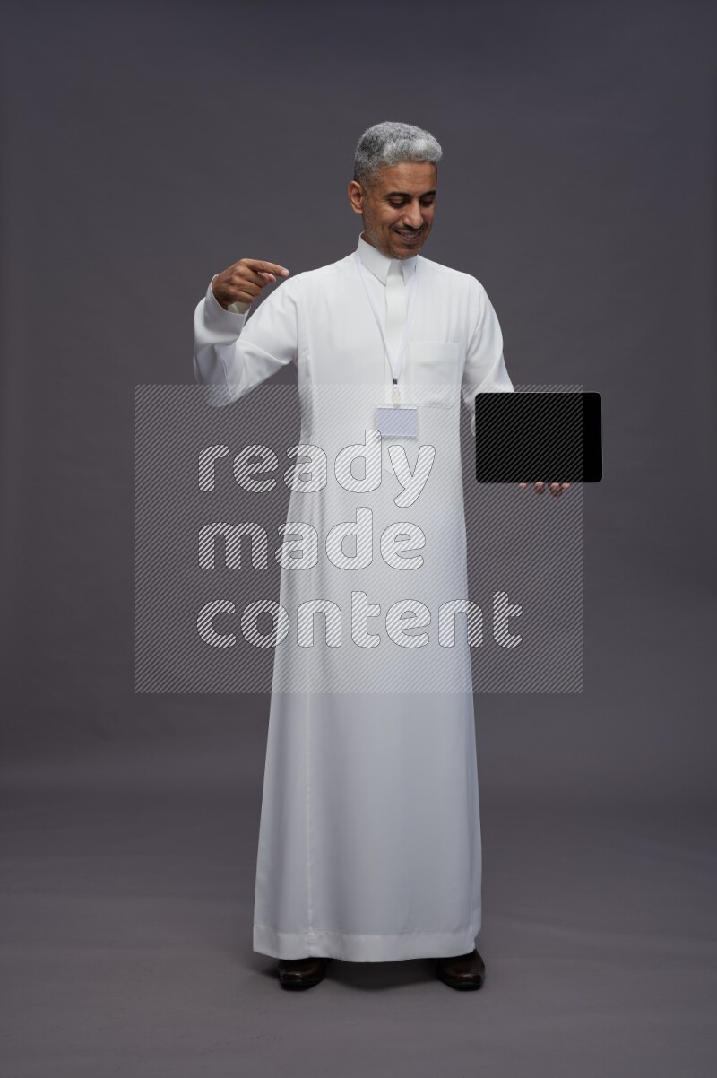 Saudi man wearing thob with neck strap employee badge standing showing tablet to camera on gray background