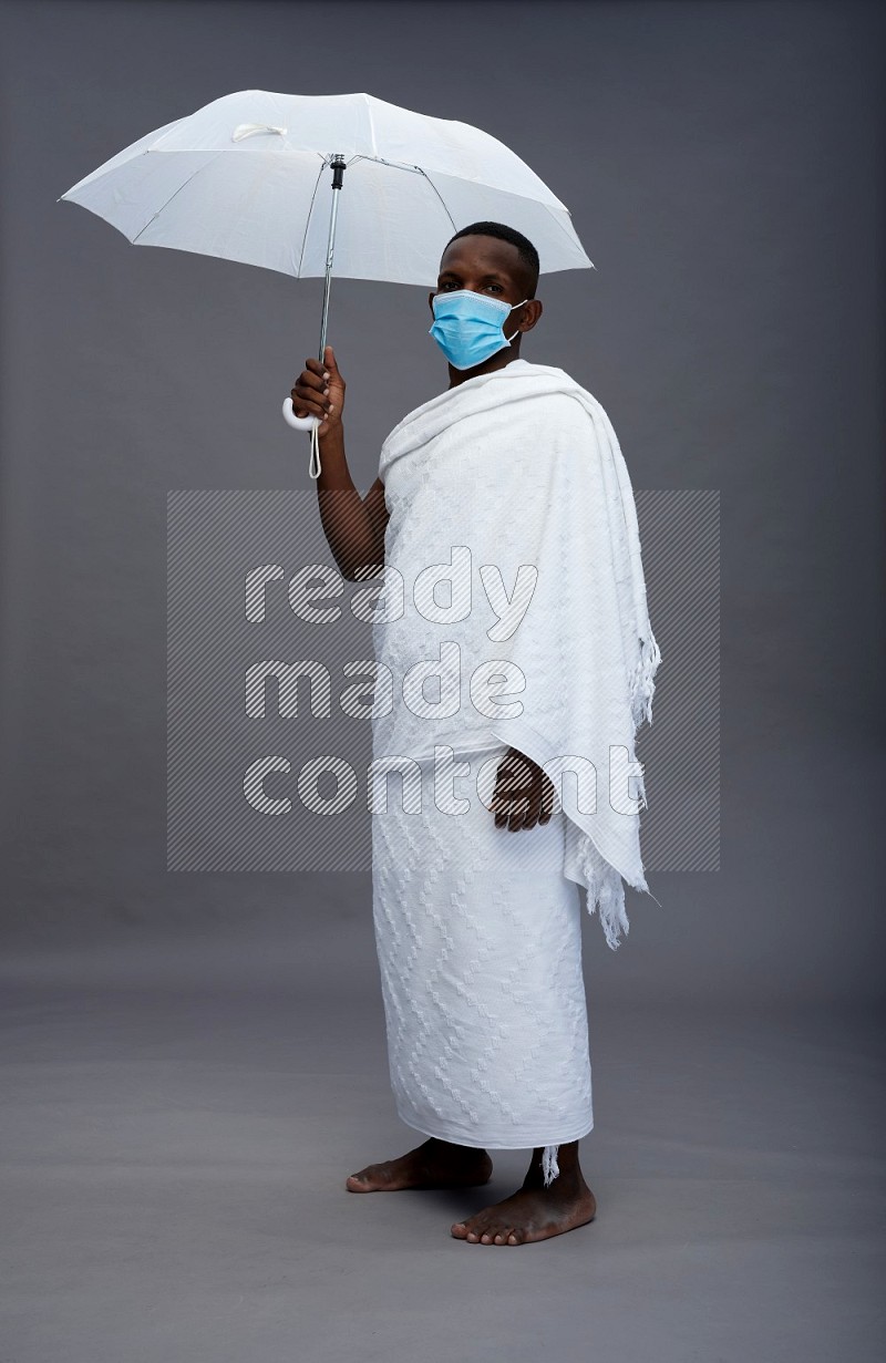 A man wearing Ehram with face mask Standing holding umbrella on gray background