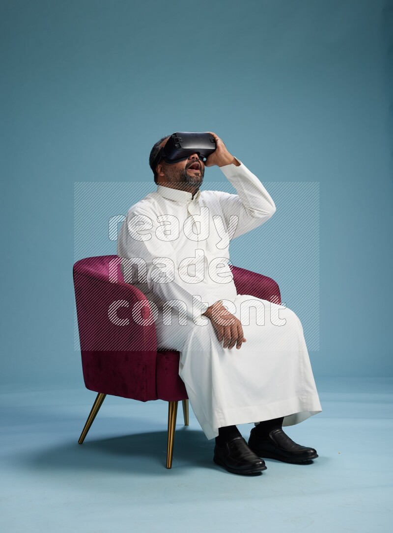 Saudi Man without shimag sitting on chair  gaming with VR on blue background
