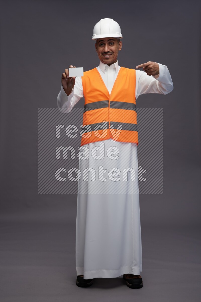 Saudi man wearing thob with engineer vest standing holding ATM card on gray background