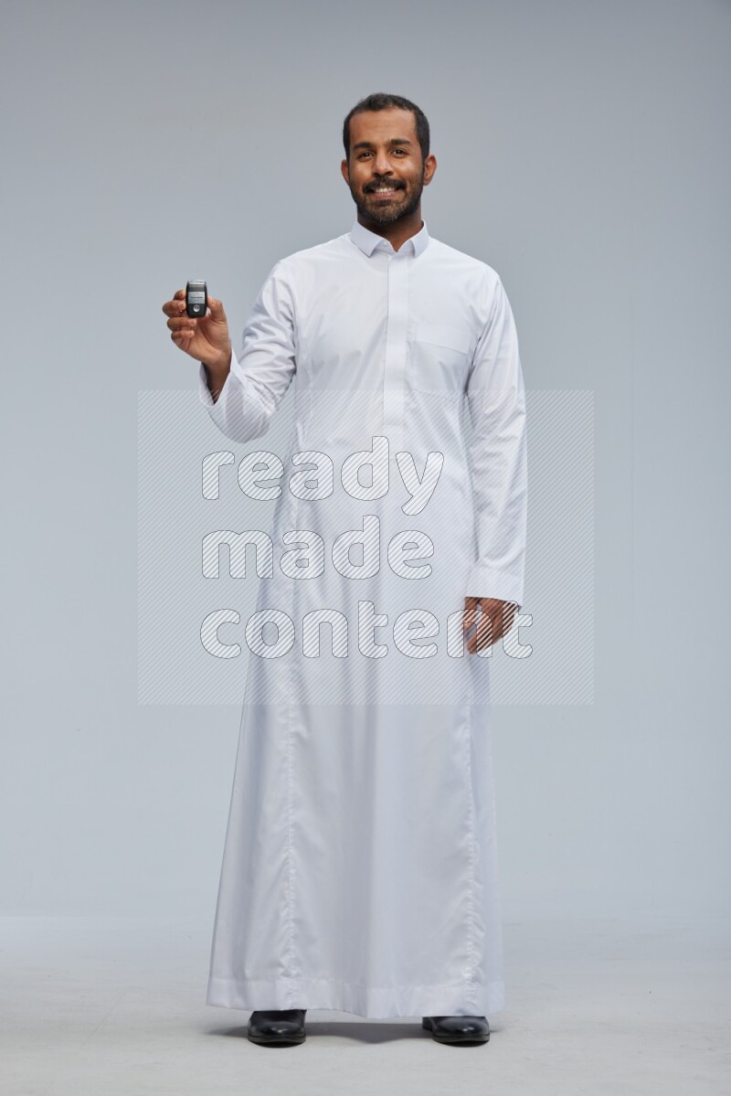 Saudi man wearing thob and shomag standing holding car key on gray background