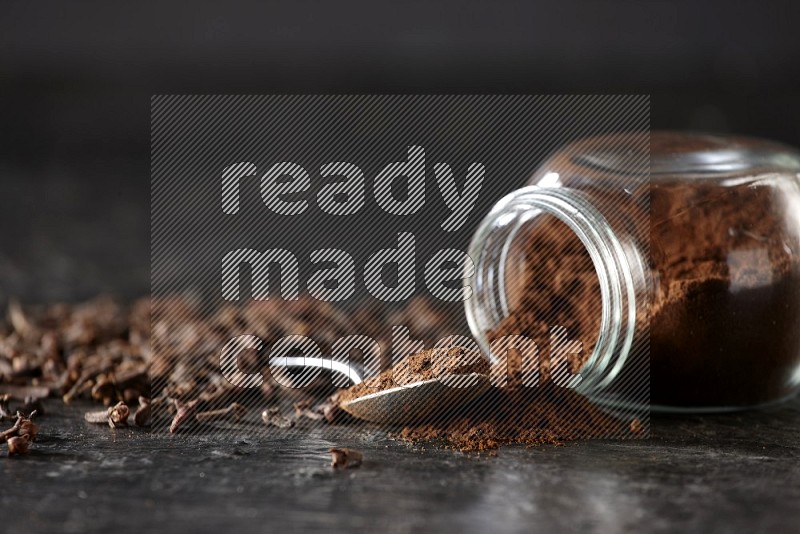 A flipped glass spice jar and a metal spoon full of cloves powder with cloves spread on textured black flooring
