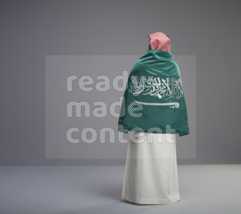 A Saudi man standing wearing thob and red shomag wrapping big saudi flag on gray background