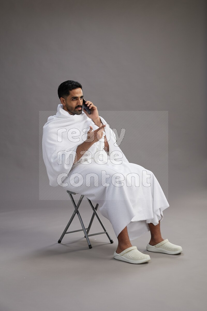 A man wearing Ehram sitting on chair talking on the phone on gray background