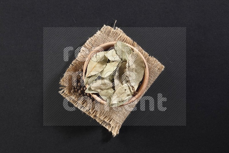 A wooden bowl filled with laurel bay on a piece of burlap with some of allspice berries on black flooring in different angles