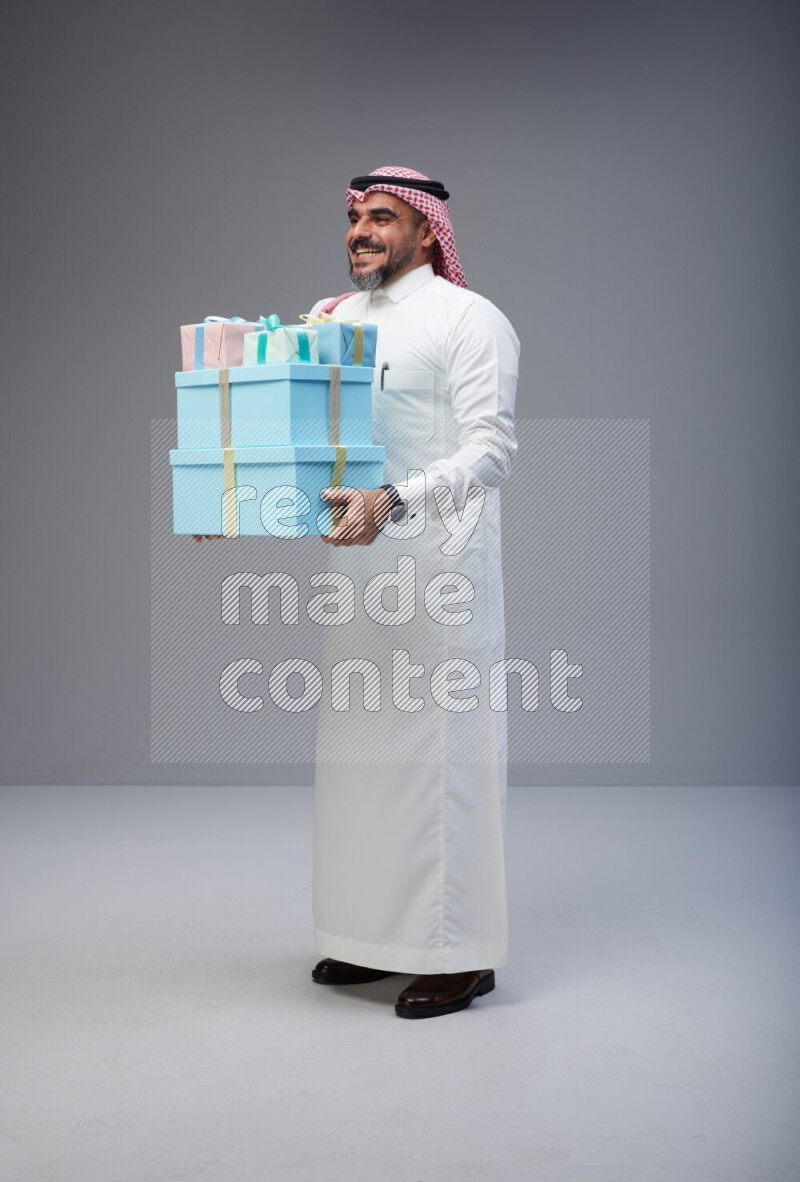 Saudi man Wearing Thob and red Shomag standing holding gift box on Gray background