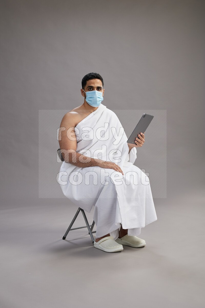 A man wearing Ehram with face mask sitting on chair holding a tablet on gray background
