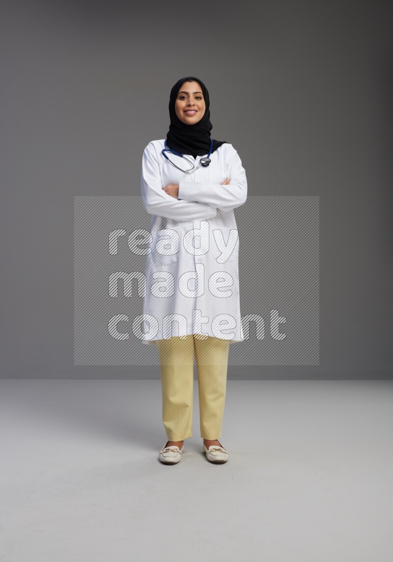Saudi woman wearing lab coat with stethoscope standing with crossed arms on Gray background