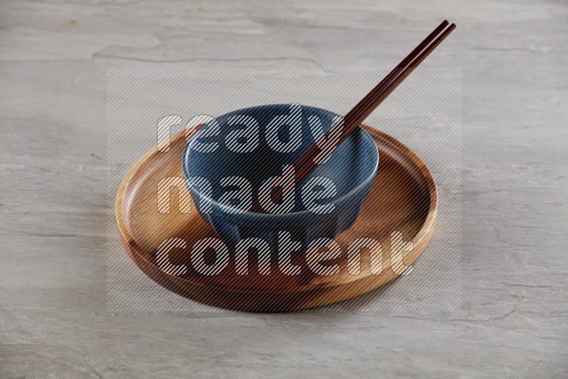 Blue pottery round bowl on top of brown wood round plate and wood chopsticks, on grey textured countertop