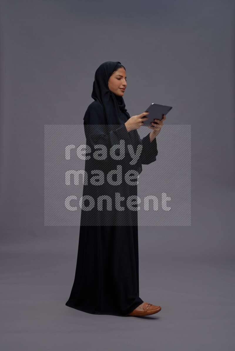 Saudi woman wearing Abaya standing working on tablet on gray background