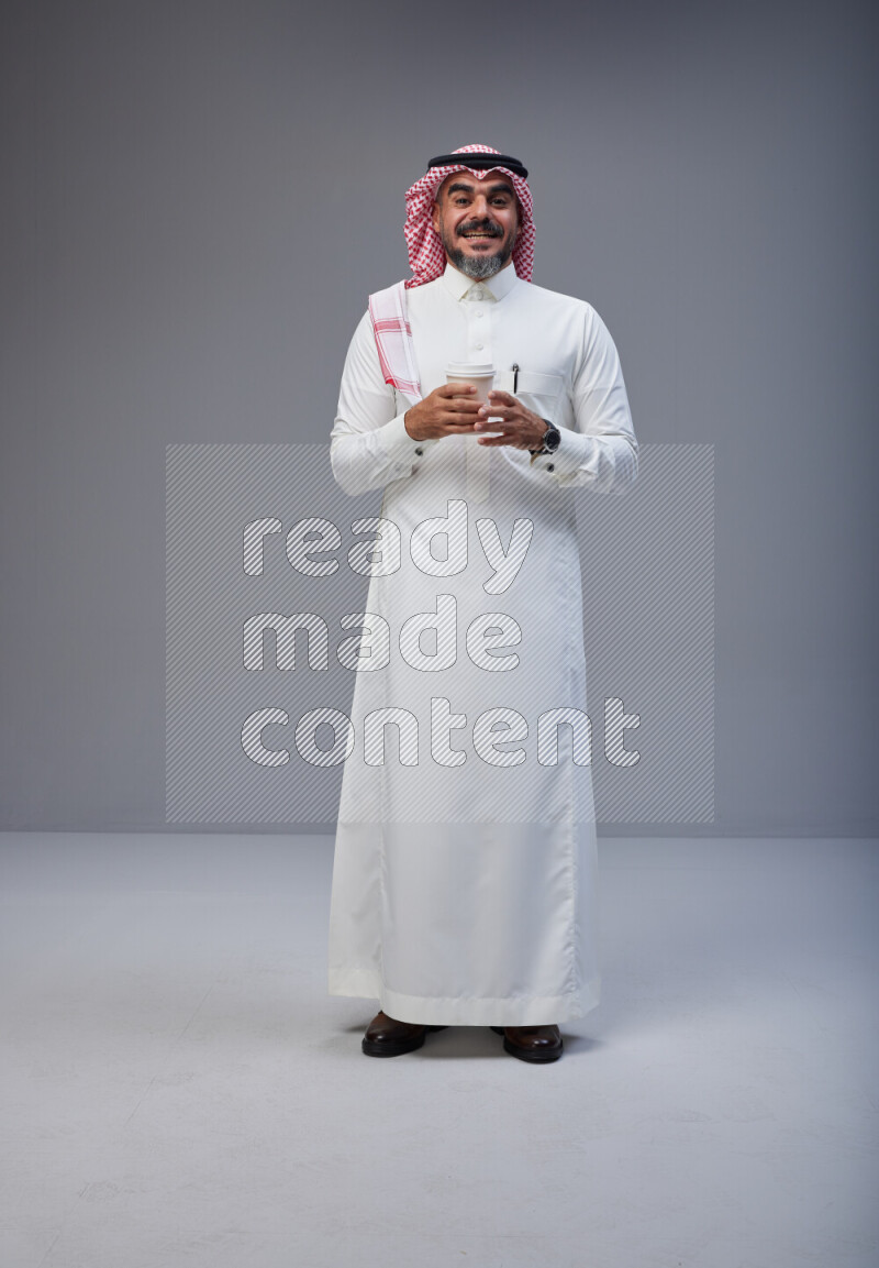 Saudi man Wearing Thob and red Shomag standing holding paper cup on Gray background