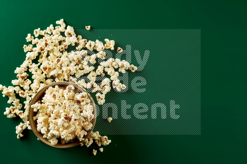 An off white ceramic bowl full of popcorn with popcorn beside it on a green background in a top view shot