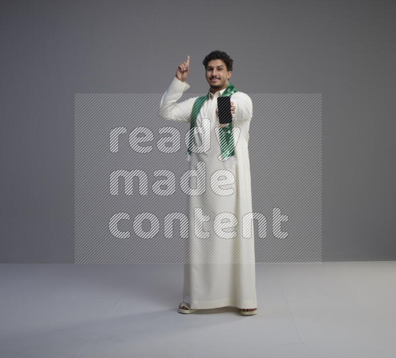A Saudi man standing wearing thob and Saudi flag scarf showing phone to camera on gray background