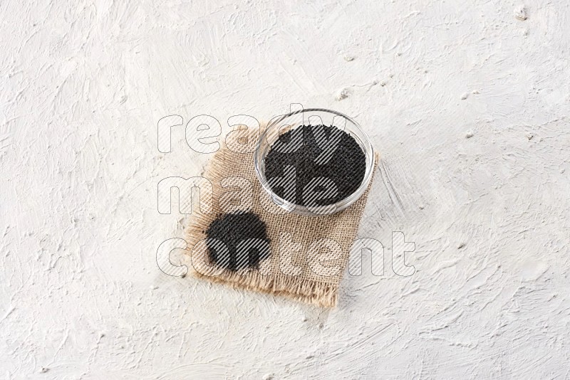 A glass bowl full of black seeds on a burlap piece on textured white flooring