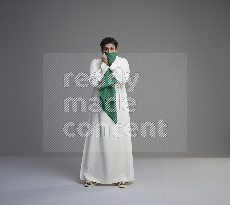 A Saudi man standing wearing thob kissing Saudi flag on gray background