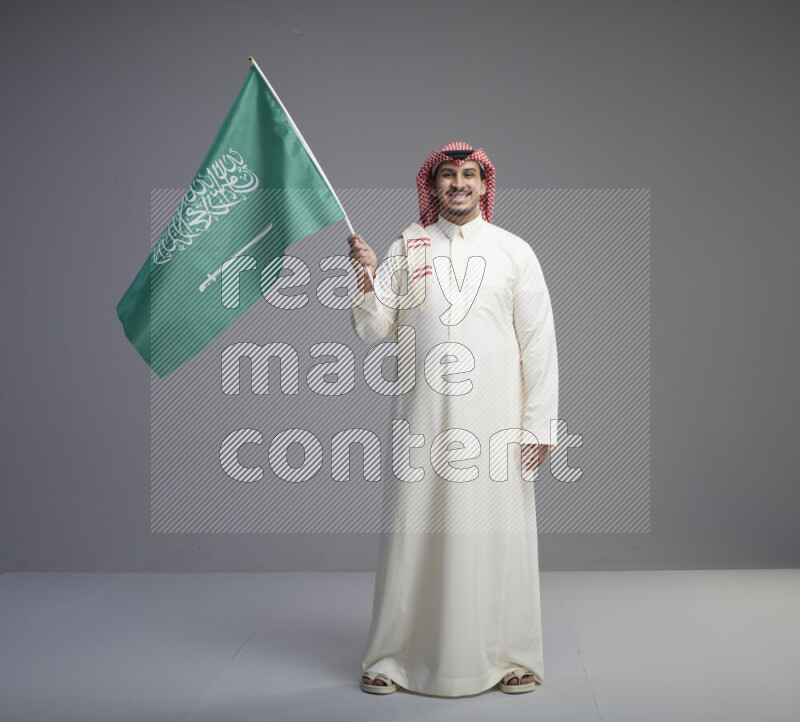 A Saudi man standing wearing thob and red shomag raising big Saudi flag on gray background
