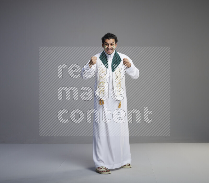 A Saudi man standing wearing thob and Saudi flag scarf with face painting on gray background