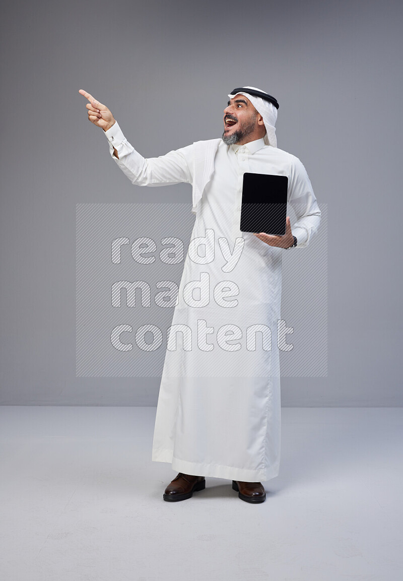 Saudi man Wearing Thob and white Shomag standing showing tablet to camera on Gray background