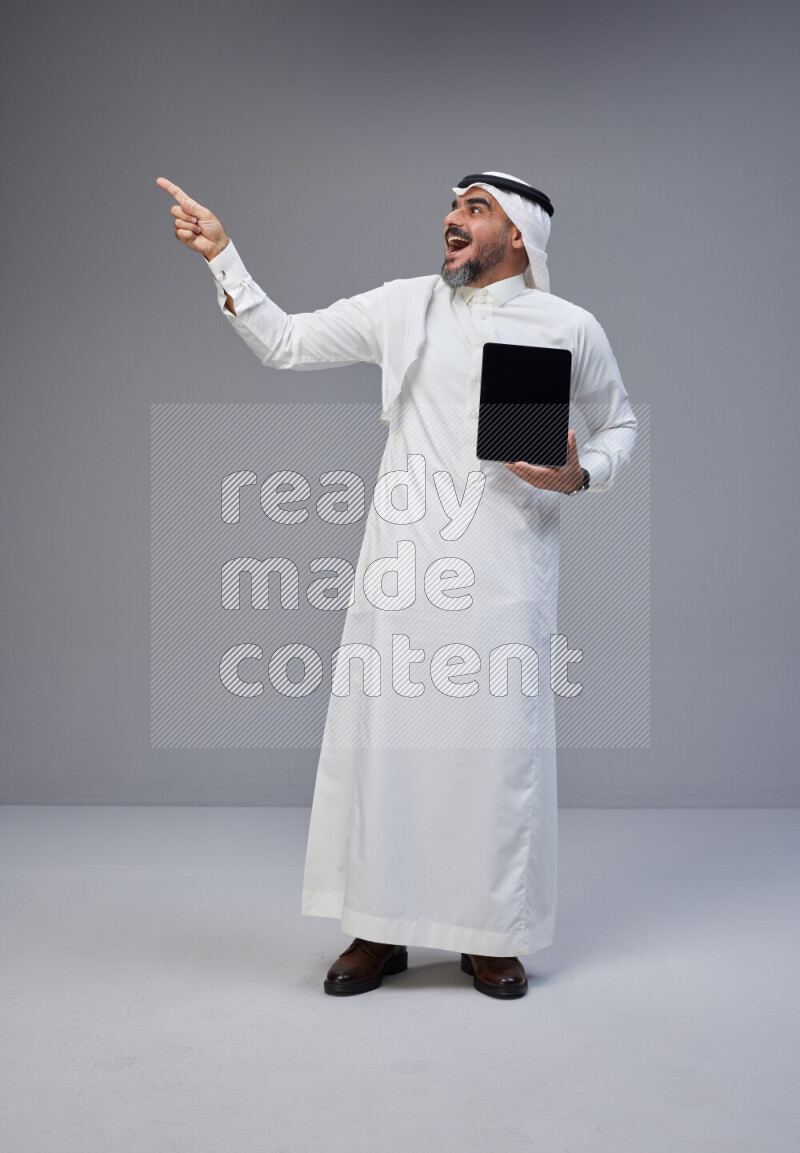 Saudi man Wearing Thob and white Shomag standing showing tablet to camera on Gray background