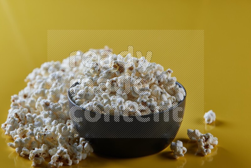 A black ceramic bowl full of popcorn with popcorn beside it on a yellow background in different angles