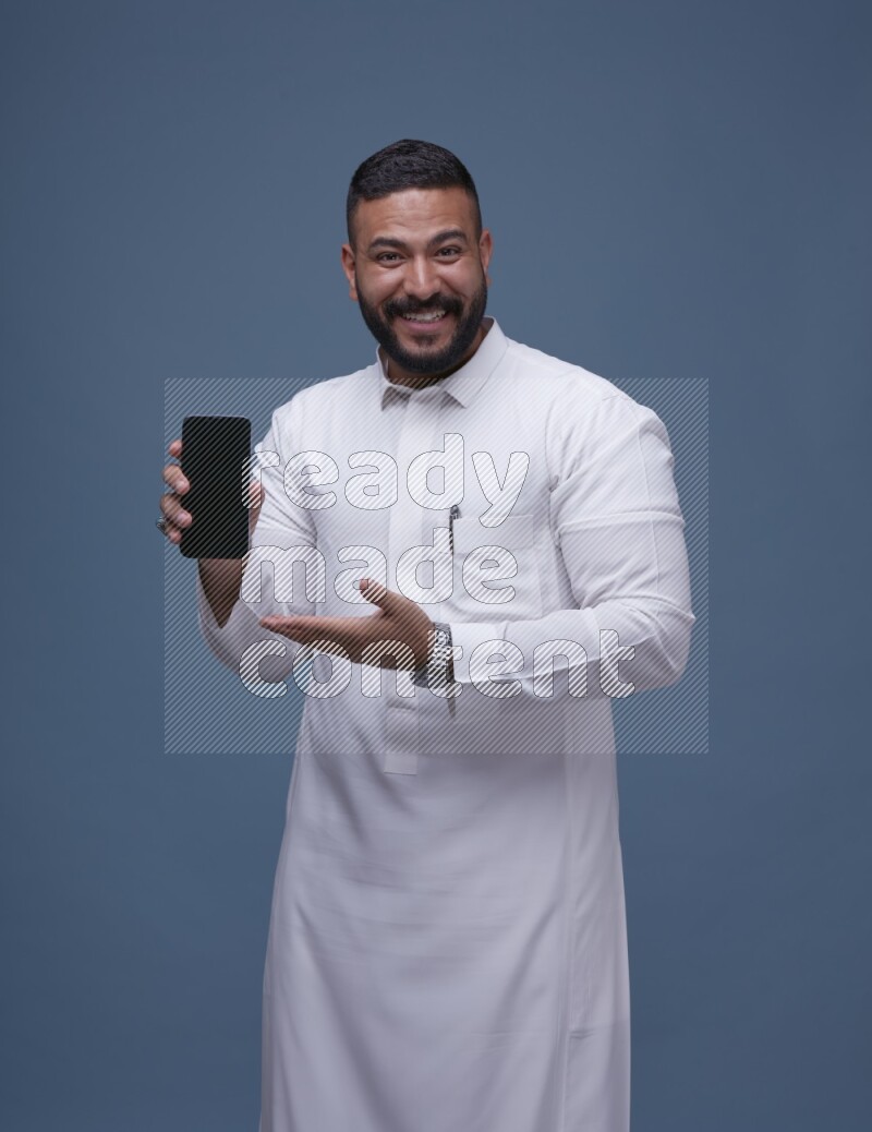 A man Showing his phone in Blue Background wearing Saudi Thob