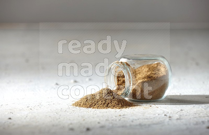 A flipped glass spice jar full of cumin powder and powder spilled out on textured white flooring