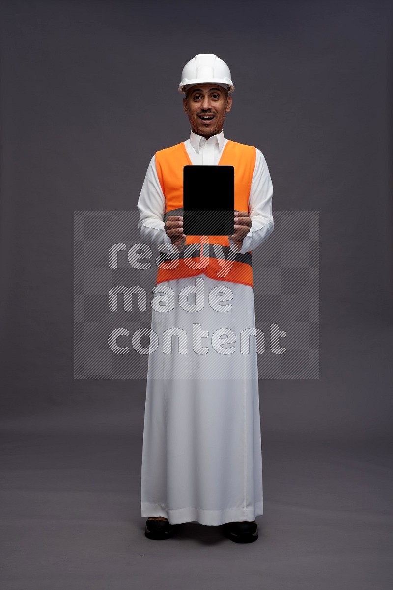 Saudi man wearing thob with engineer vest standing showing tablet to camera on gray background