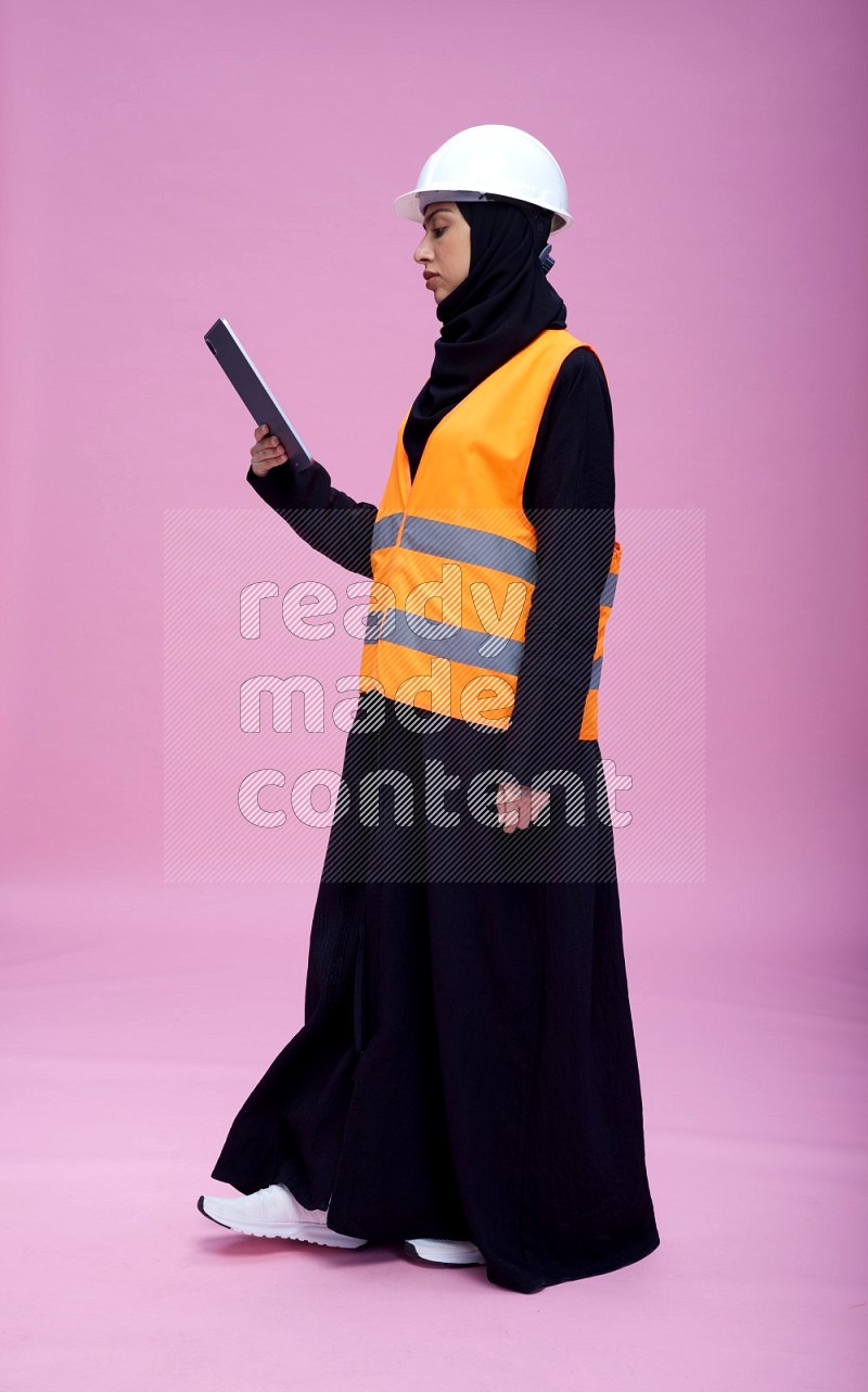 Saudi woman wearing Abaya with engineer vest and helmet standing working on tablet on pink background