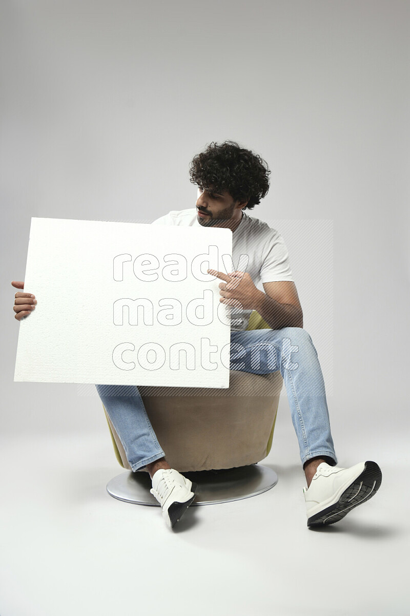 رجل يرتدي ملابس كاجوال يجلس علي كرسي و يحمل لوحة بيضاء علي خلفية بيضاء