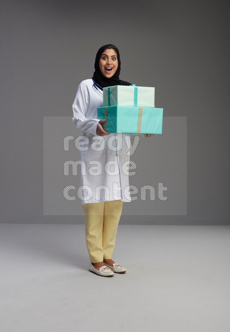 Saudi woman wearing lab coat with stethoscope standing holding gift box on Gray background