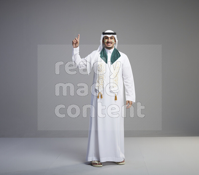 A Saudi man standing wearing thob and white shomag with face painting and Saudi flag scarf on gray background