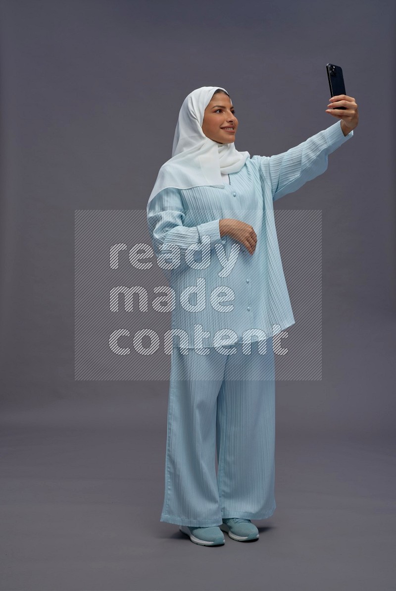 Saudi woman wearing hijab clothes standing taking selfie on gray background