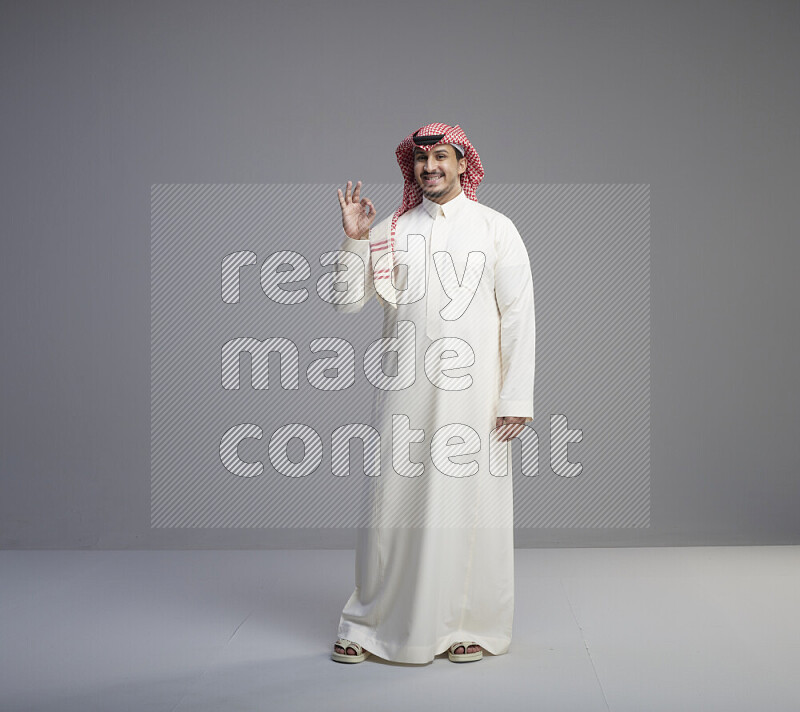 A Saudi man standing wearing thob and red shomag interacting with the camera on gray background