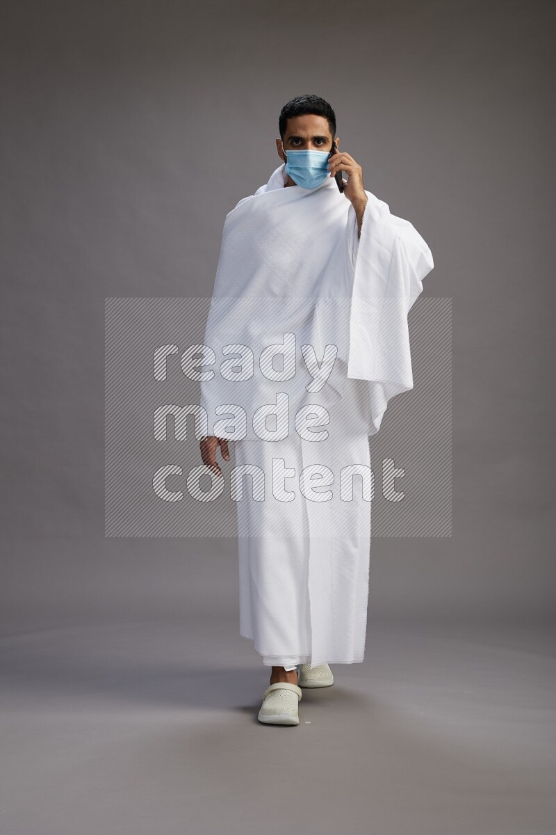 A man wearing Ehram with face mask Standing talking on phone on gray background